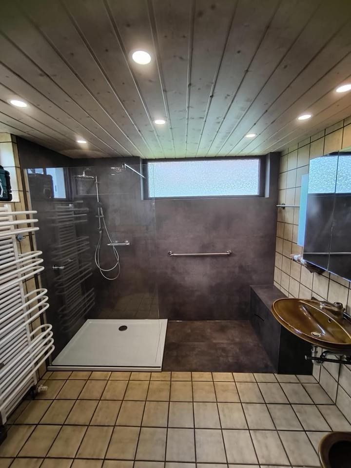 Nachher: dasselbe Bad mit großer Dusche, dunklen Wandpaneelen und Haltegriff