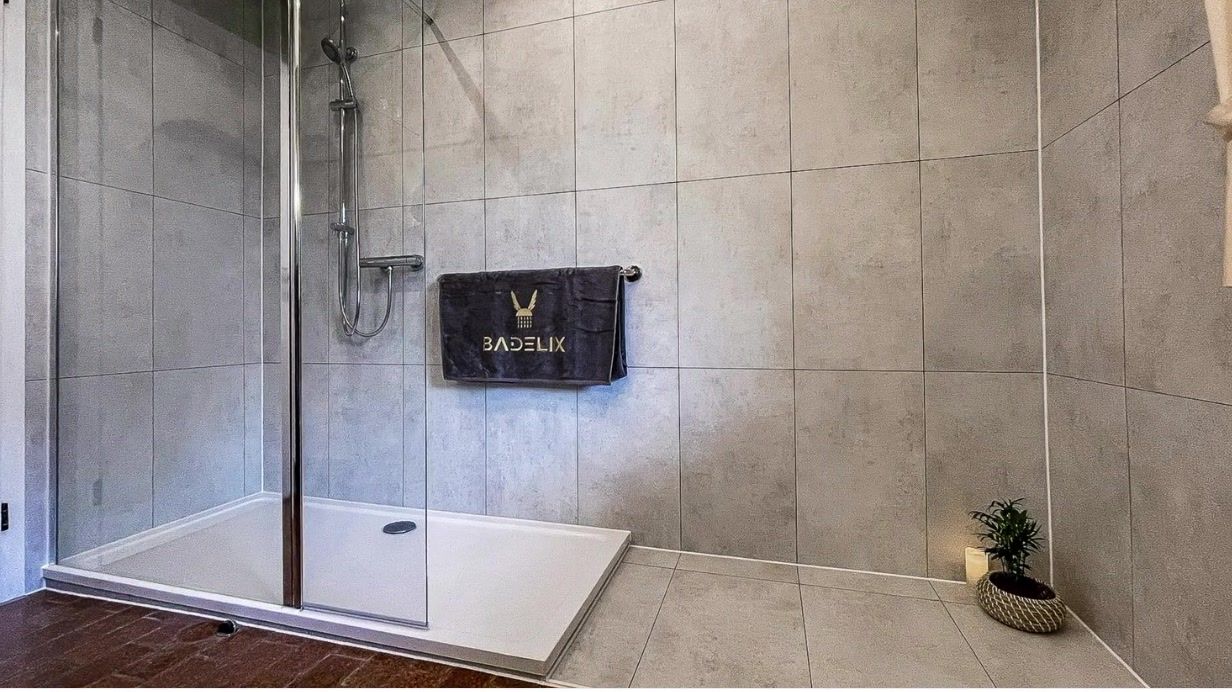 Fertige Dusche aus einem echten Badelix-Umbau – graue Wandpaneele, Glaswand, Handtuch mit Badelix-Logo